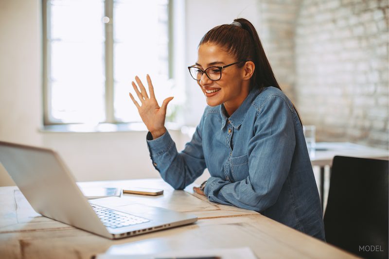 Woman seated in front of computer, waving to others on a video conference.