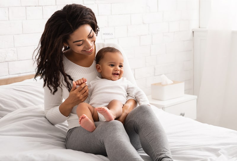 African American woman with her young baby sitting on her lap on bed.