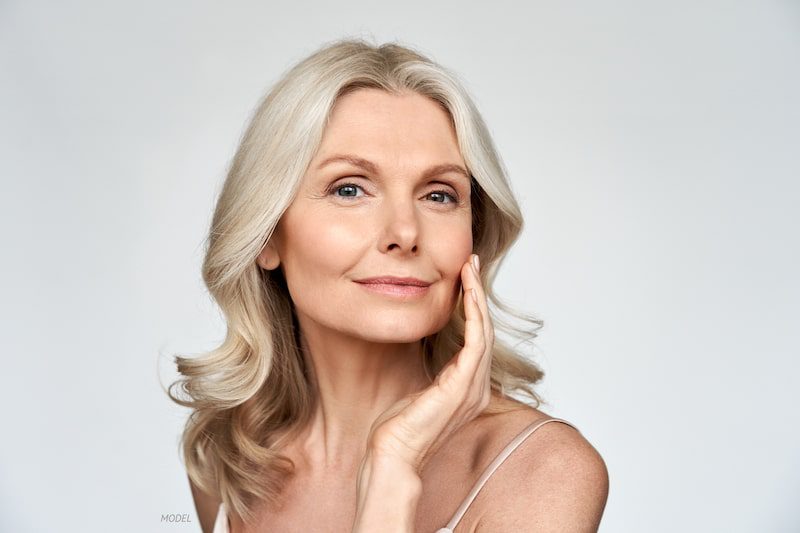 Middle-aged woman looking content as she touches her face against a gray background.