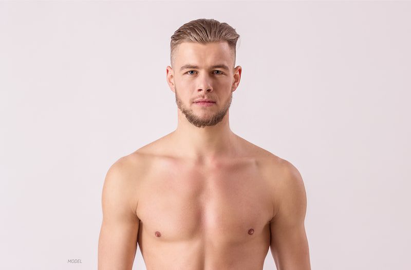 Shirtless young man with short beard standing against a tan background.