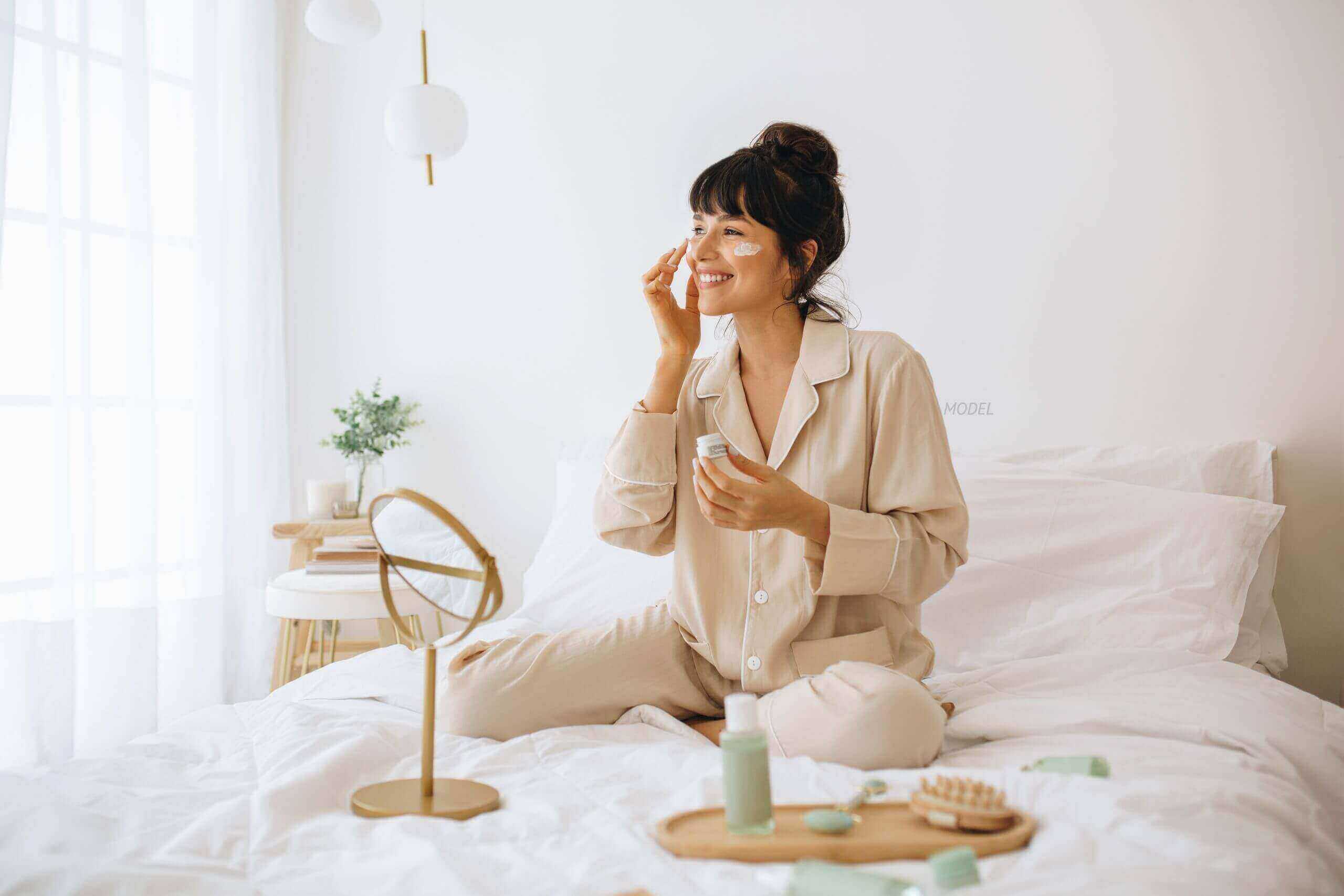 Woman applying skin care cream to cheeks while sitting on bed.