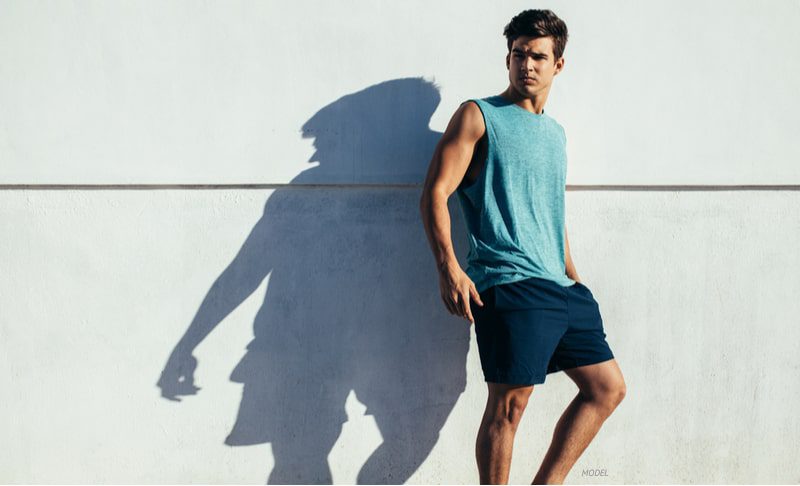 Fit man, wearing a tank-top and shorts and leaning up against a wall