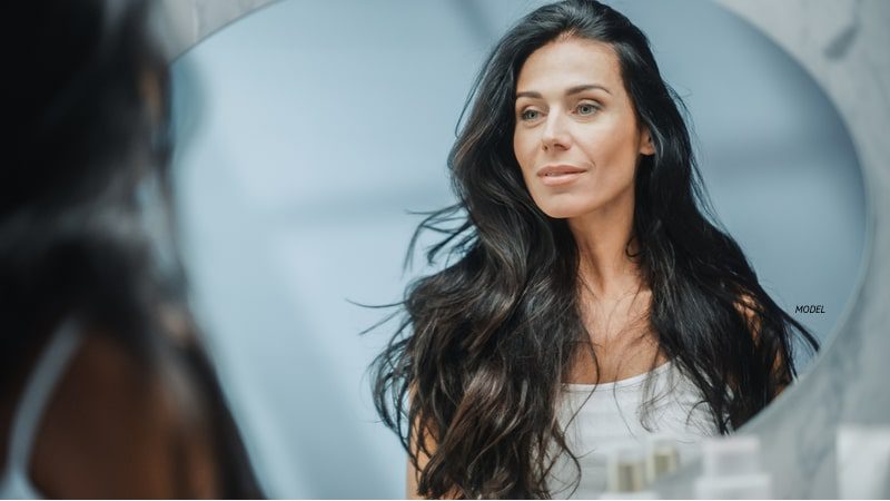 Middle-aged woman with long brunette hair looking in mirror.