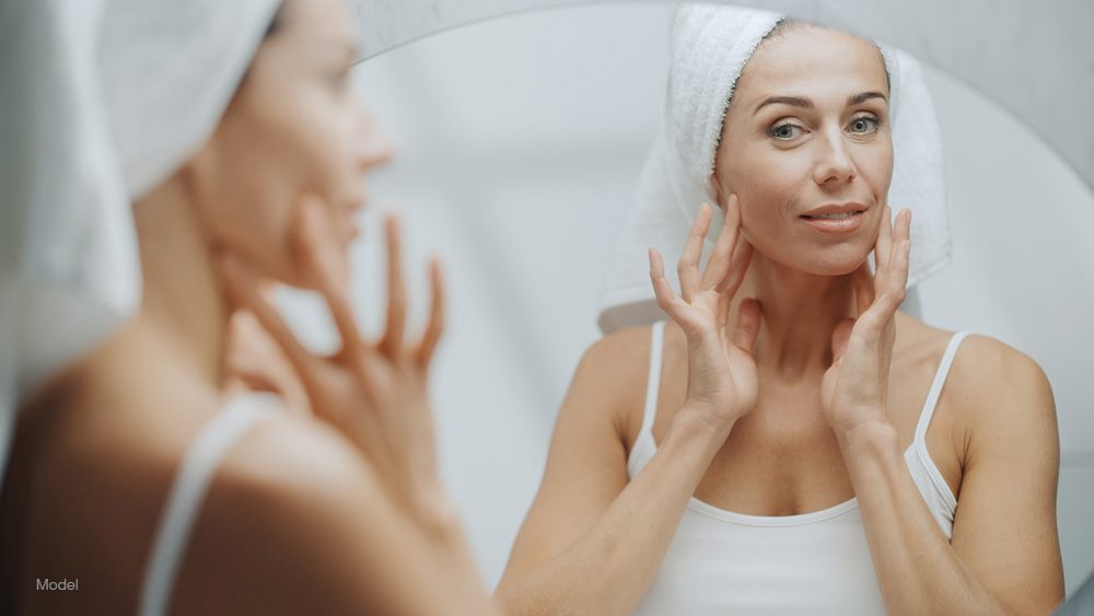 Beautiful middle-aged woman looks at her reflection in the mirror while touching the sides of her face